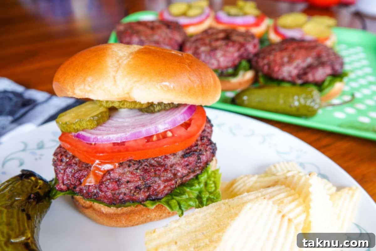 Smoked burger with fresh lettuce, tomato, and onion on a plate, with more burgers on a platter in the background.