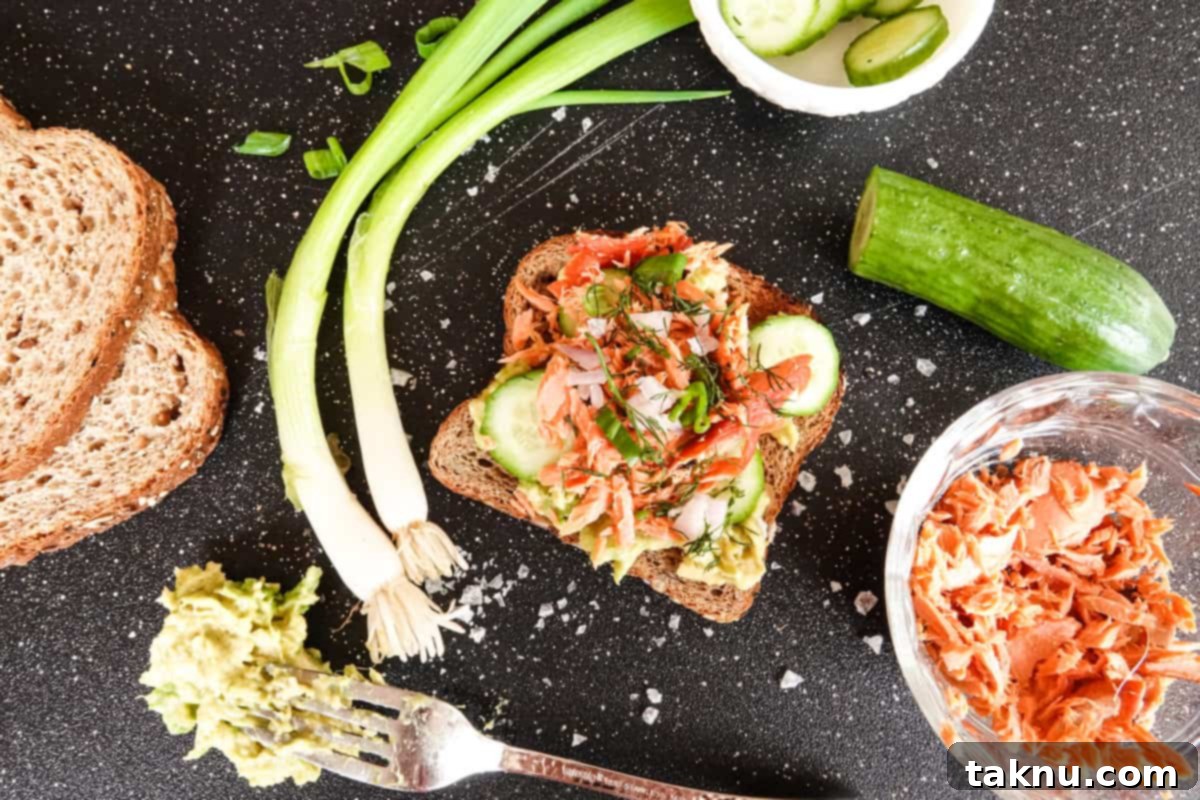 Elegant serving of salmon avocado toast alongside a bowl of flaked smoked salmon, cucumber, bread, and green onions on a black cutting board