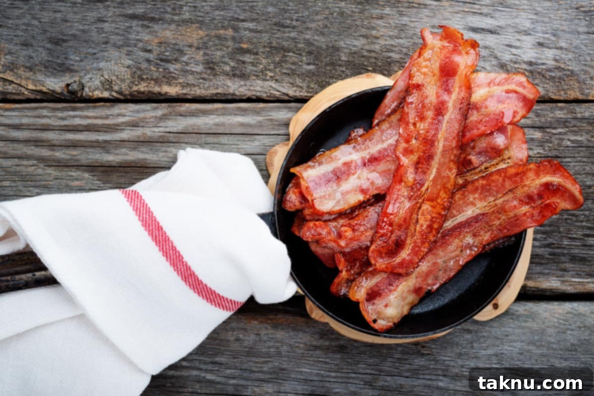 Crispy bacon sizzling in a frying pan on a stovetop, with a paper towel nearby for draining.