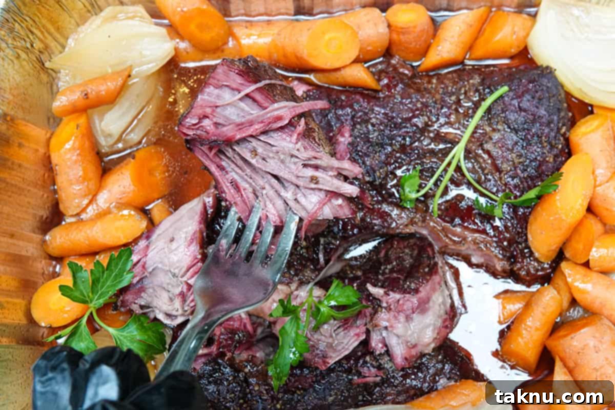 Tender beef chuck roast pulled apart with a fork, surrounded by soft carrots and onions in a serving dish.