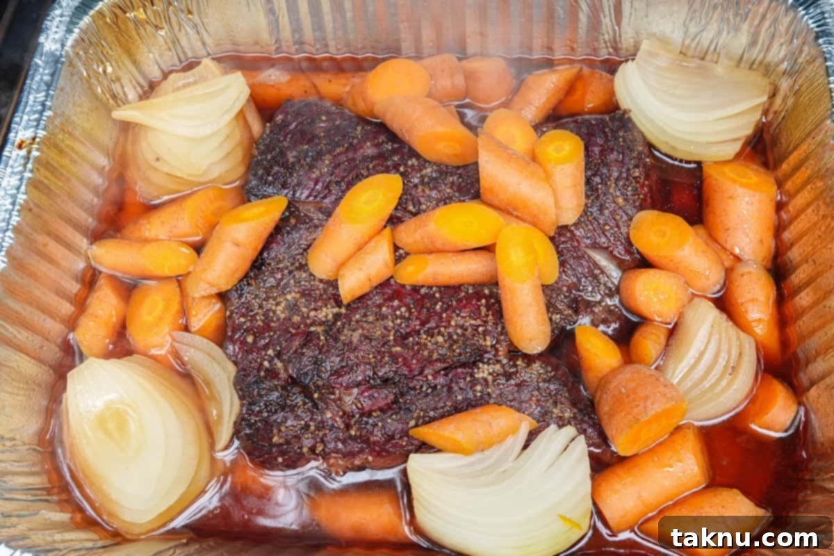 Smoked chuck roast in an aluminum pan with beef broth, quartered onions, and carrots, ready to be covered.