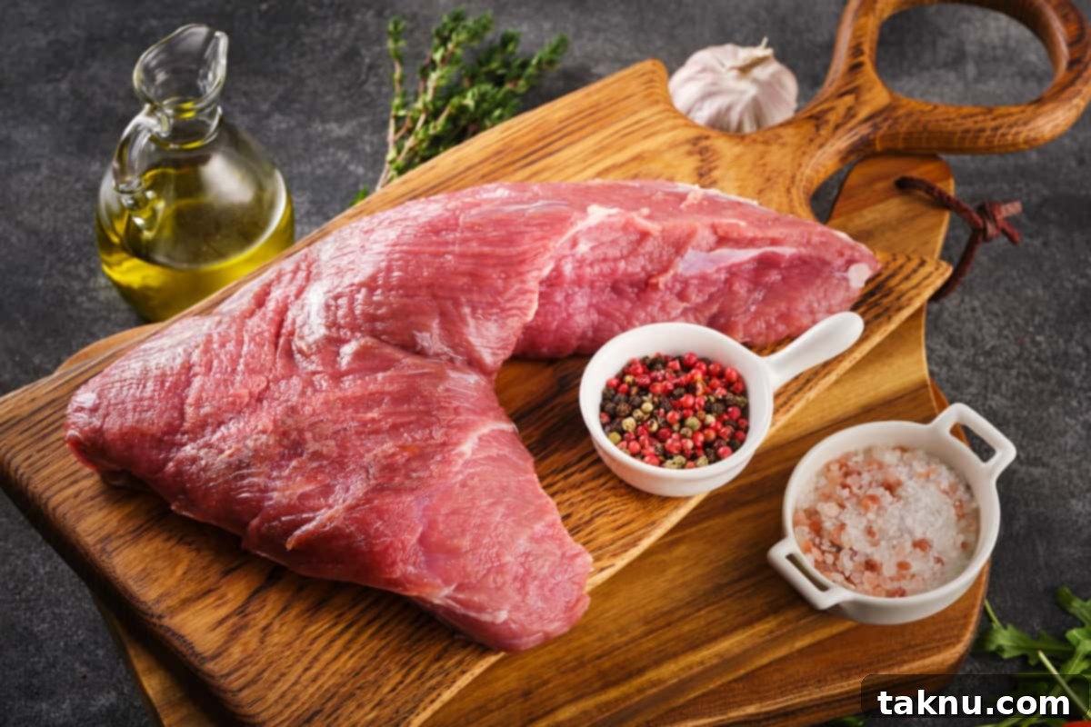 Uncooked tri-tip steak on a cutting board, surrounded by coarse salt, black pepper, and olive oil for seasoning.