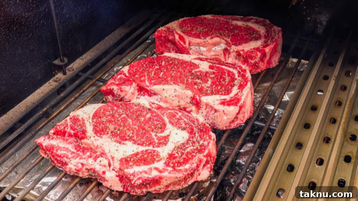 Ultimate Smoked Ribeye 5 Three seasoned ribeye steaks perfectly placed on the grates of a smoker, undergoing the low-and-slow smoking process.